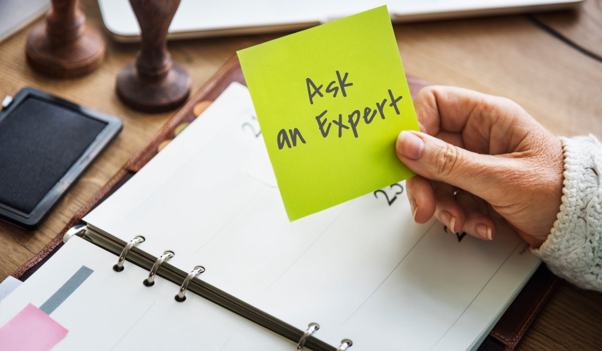 A hand holding a post-it-note with "Ask An expert" written on it. There's a calendar book on the table, a stamp and a smartphone