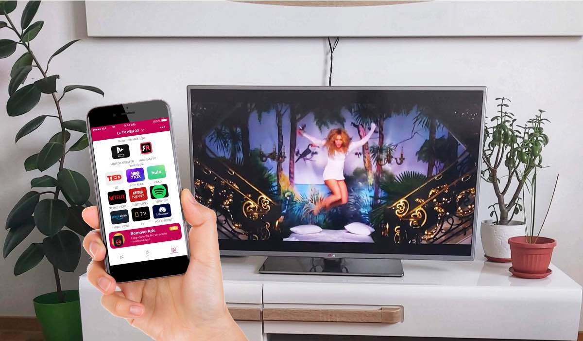 A hand holding an iPhone with the LG Tv Remote Control Plus App Launcher feature on the screen. An LG TV on a drawer with plants around