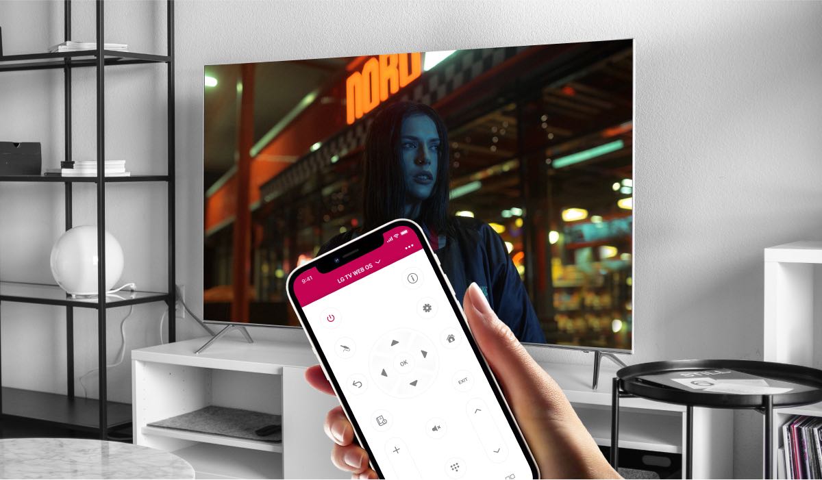 A hand holding an iPhone with LG TV Remote Control Plus app on the screen. The iPhone is pointing at a TV on a drawer. The Tv screen displays an image of a woman standing in neon-lit city