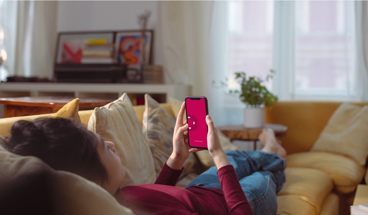 A woman laying on a couch in a living room. She's holding an iPhone with the LG TV Remote Control Plus logo on the screen