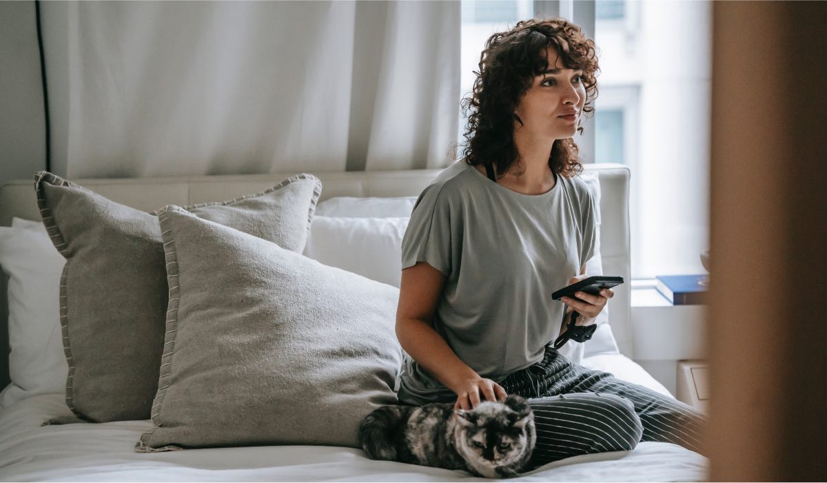 A woman sitting on a bed with pillows behind her and a sunny window on her left. She's holding a smartphone.