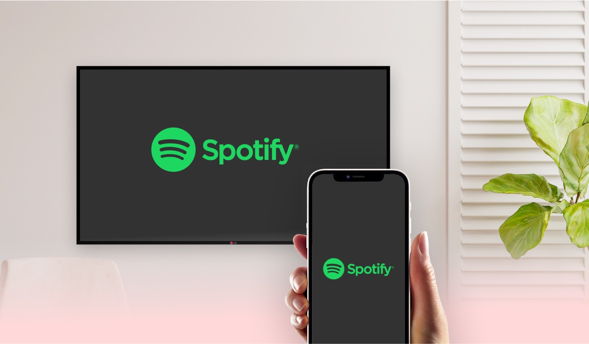 A hand holding an iPhone with Spotify logo on the screen. A smart TV with Spotify logo on the screen, a curtain and a house plant
