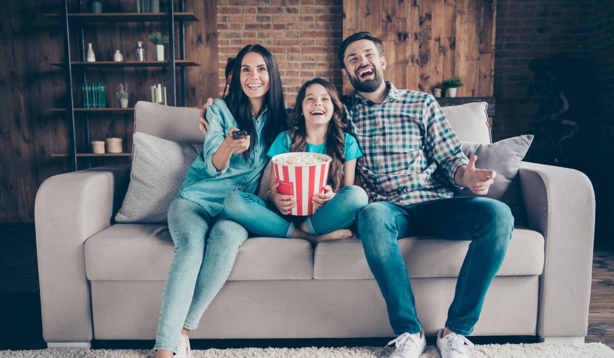 Mom, dad and daughter sit on a couch with a cinema-style container of popccorn