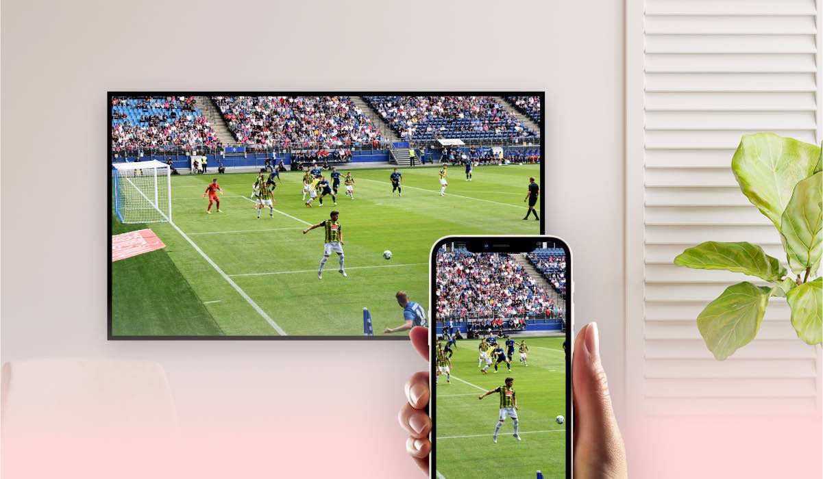 A hand holding an iPhone that is mirroring an american football match to an LG TV that is hanging on a wall, next to the curtains and a plant