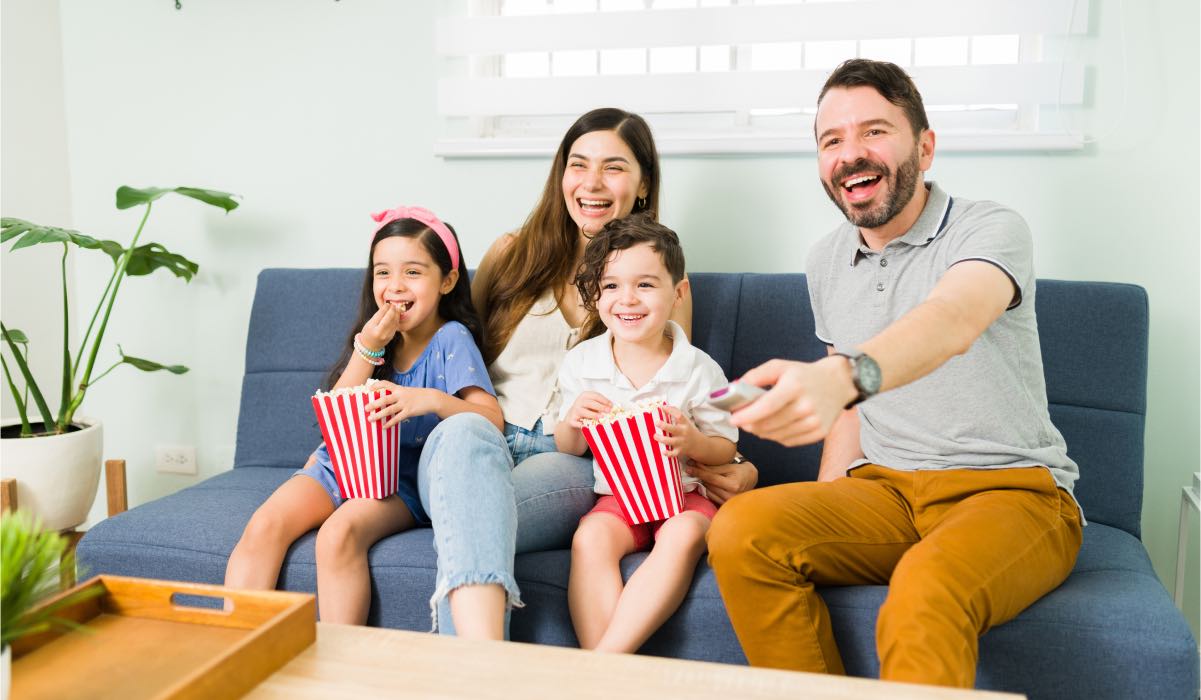 Two kids and their parents sit on a couch. The father is holding a remote control. The kids hold popcorn buckets