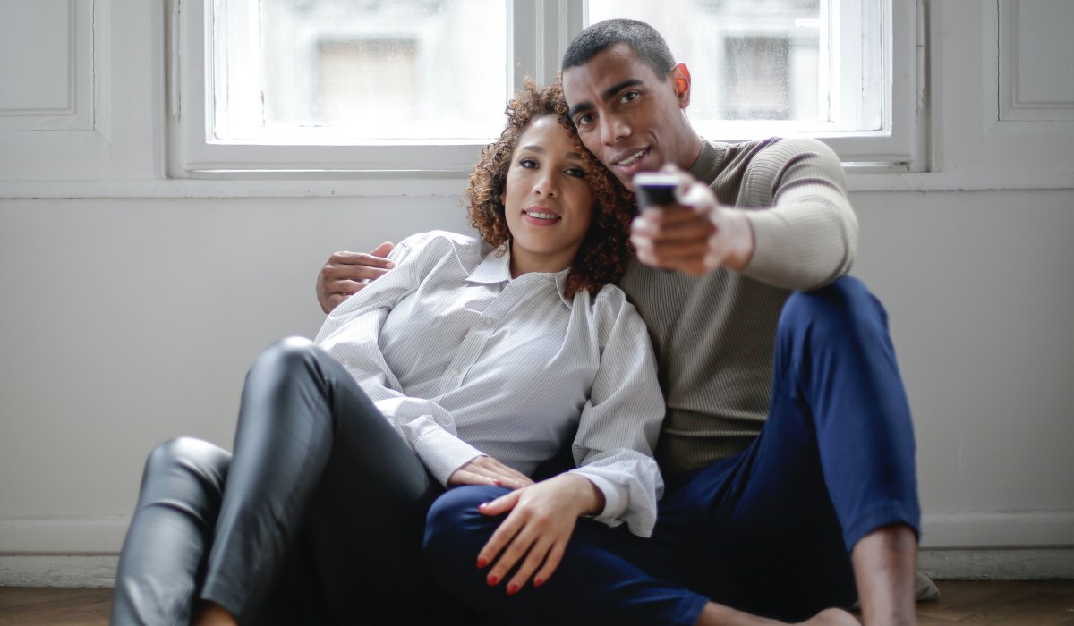 A man and a woman sit in an embrace. The man is pointing a remote control in front of him