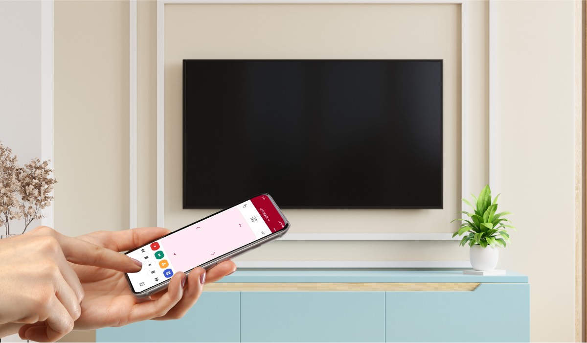 A hand holding an iPhone with the LG TV Remote Control Plus app interface on the screen. An LG TV hanging on a wall. It has a black screen