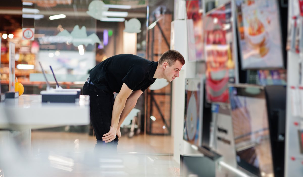 A man in an electronics store, bending down to see a TV