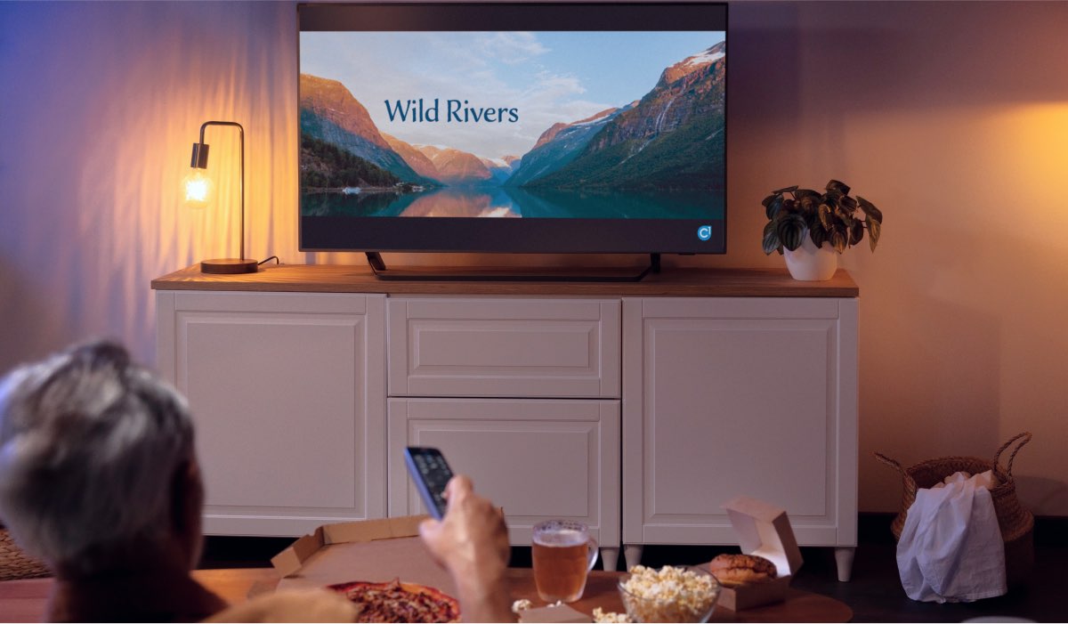 A person holding a remote, pointing it at an LG TV on a white drawer. The TV has "Wild Rivers" on the screen