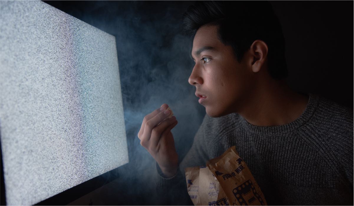 A man watching TV closely to the screen with a bowl of popcorn. The TV displays TV static - the light illuminates the man's face