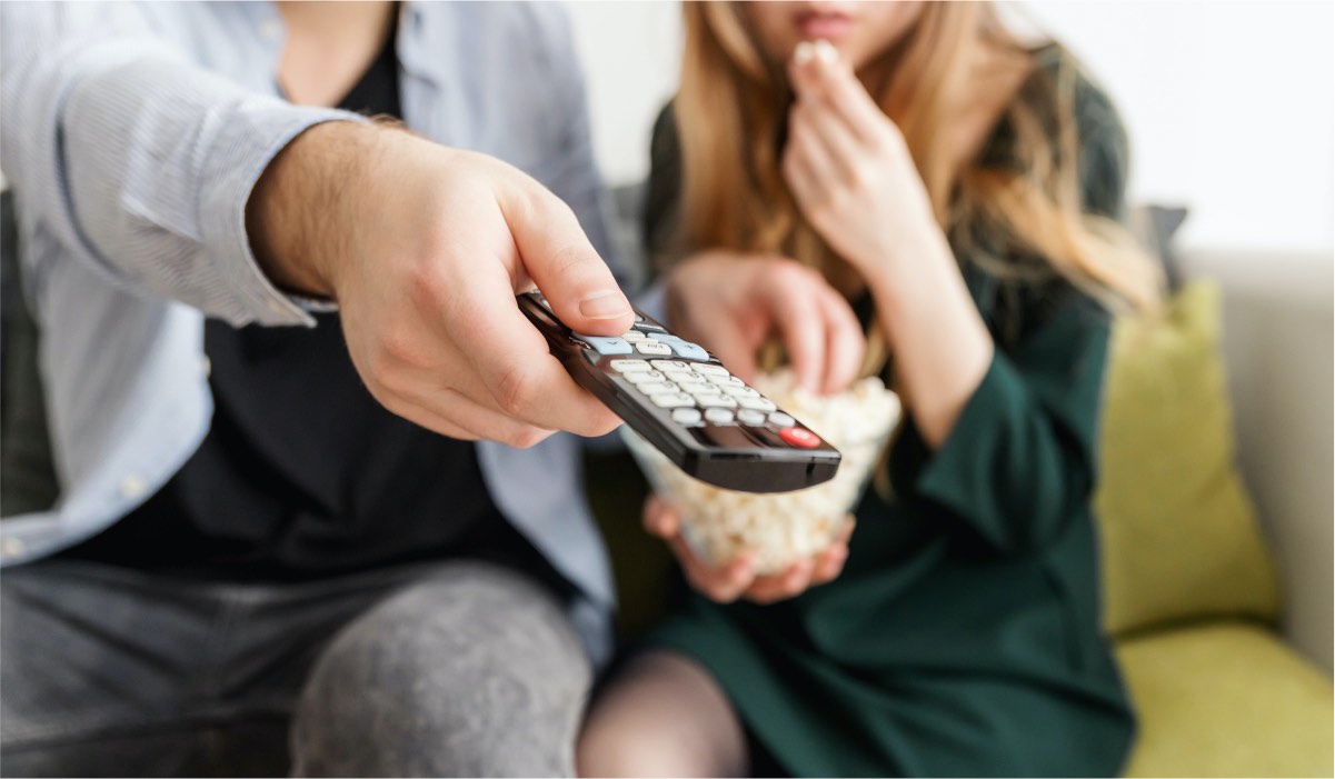 A person holding a remote and another person holding a bowl of popcorn. They are both sitting on a couch