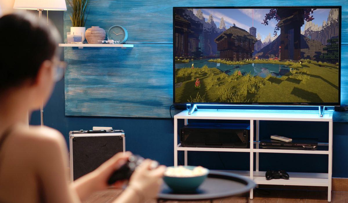 A woman playing Minecraft on a console plugged to an LG TV. The TV is standing on a white drawer and there's a coffee table with snacks