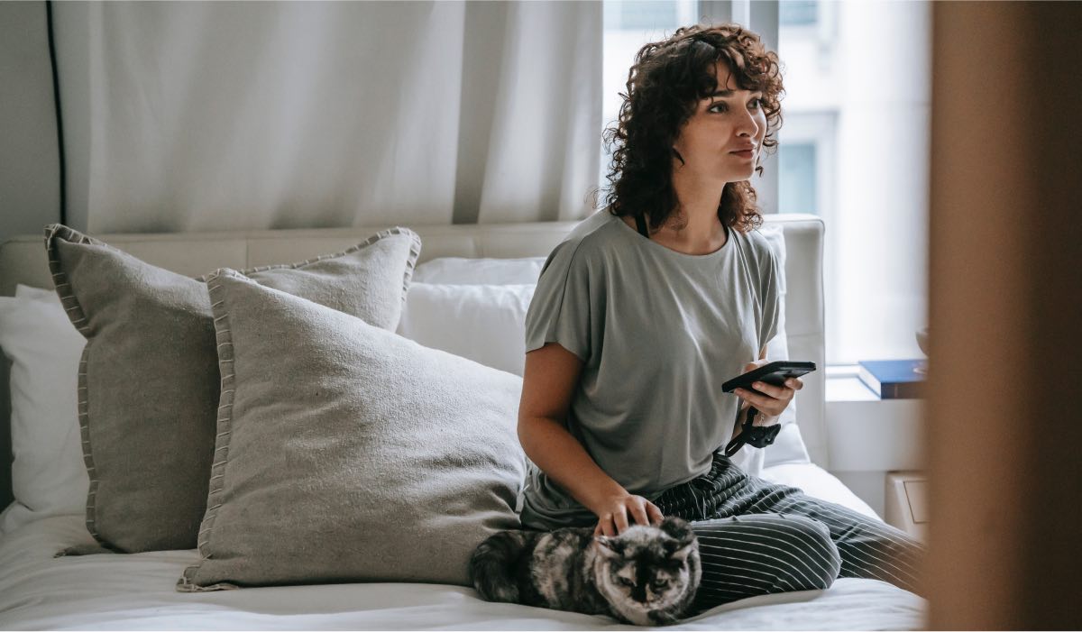 A woman sitting on her bed, looking at a TV. She is holding a smartphone in one hand, her second hand is stroking a cat that is laying beside her.