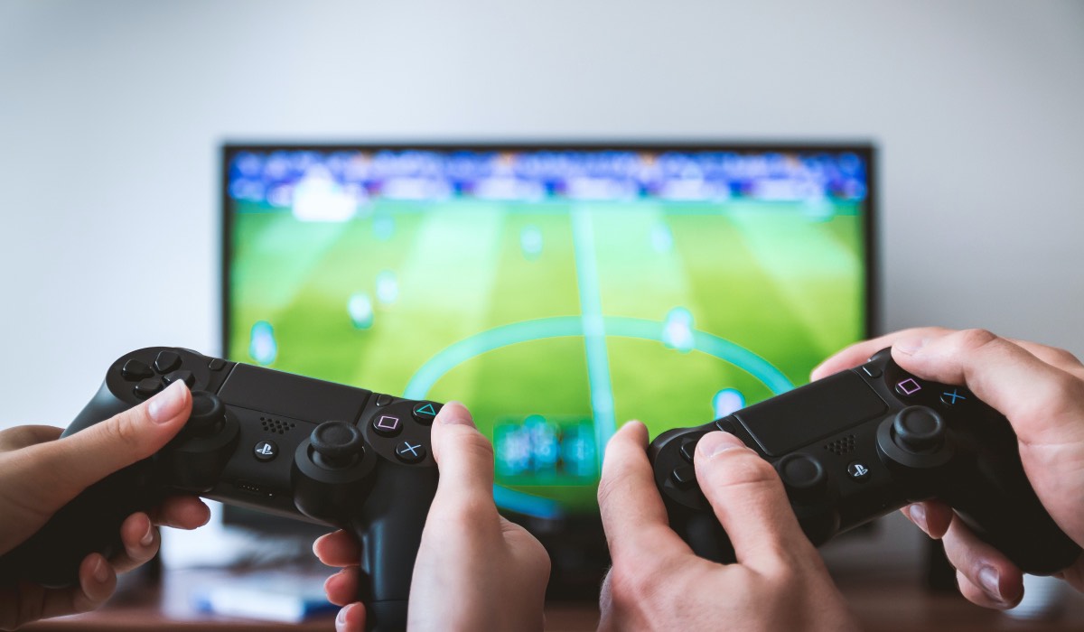 Two sets of hands holding gaming console controllers. A football video game on a TV